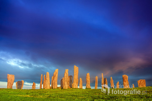 Fotografia przedstawia zespół stojacych głazów o nazwie Callanish Stones, zlokalizowany na wyspie Lewis w archipelagu Hebrydów Zewnętrzych administracyjnie należącego do Szkocji. Głazy stanowią formę starożytnej, celtyckiej świątyni. Był to jeden z głównych celów mojej ówczesnej wędrówki po wybrzeżu szkockim.
Pomimo gęstych chmur jakże charakterystycznych dla Szkocji, ostatnie promienie zachodzącego słońca zdołały się przebić i doświetlić owe monumentalne głazy.