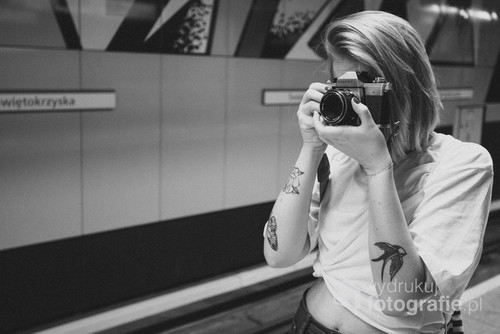 Stacja metra Świętokrzyska. Dziewczyna z analogową Prakticą, chwyta chwilę z życia miasta na klasyczną kliszę fotograficzną. Zdjęcie cyfrowe, ale dla tego tematu zastosowałem symulację kliszy fotograficznej Ilford HP5