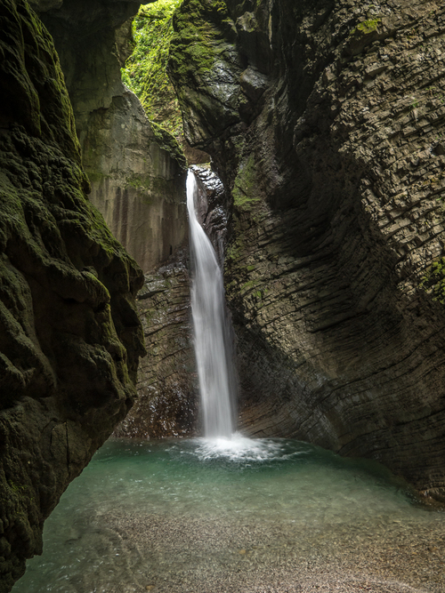 Na tym malowniczym zdjęciu ukazany jest wodospad Kozjak, jedno z przyrodniczych cudów Słowenii. Wodospad Kozjak to nie tylko imponujący przykład siły natury, ale także prawdziwa perła krajobrazu. Wodospad jest prawdziwym klejnotem przyrody.

Woda spada ze szczeliny między skałami, tworząc imponujący strumień, który następnie rozprasza się w małym akwenie, otaczając powietrze wilgocią i chłodem. Strumień wody opada na gładkie, kamieniste dno, gdzie tworzy malownicze podłoże.

Skały wokół wodospadu Kozjak dodają tej scenerii charakterystycznego uroku. Ich różne odcienie szarości i zieleni tworzą kontrast z białymi kaskadami wody, nadając krajobrazowi niezwykłą harmonię kolorów. To miejsce zachęca do kontemplacji i relaksu, a otaczająca je zieleń dodaje doświadczeniu dzikiego uroku i spokoju.

To idealne miejsce dla miłośników przyrody, turystów i fotografów, którzy chcą zachwycić się majestatyczną siłą wodospadu Kozjak oraz pięknem przyrody Słowenii. To miejsce, które z pewnością pozostawi niezapomniane wrażenie i długotrwałą pamiątkę w sercu każdego, kto odwiedził ten wspaniały zakątek natury.