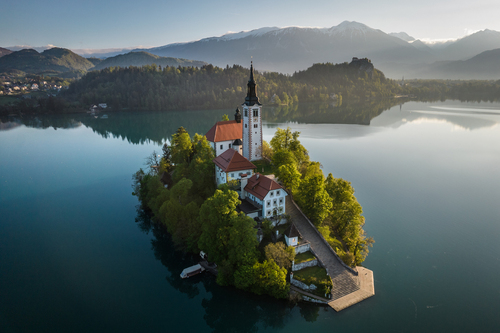 Ta malownicza wysepka, w samym sercu urokliwego Jeziora Bled, jest prawdziwym klejnotem Słowenii. Dominując na tafli spokojnej, błękitnej wody, znajduje się Cerkev Marijinega Vnebovzetja, znana jako Cerkiew Wniebowzięcia Najświętszej Maryi Panny. Ale to nie wszystko, co przyciąga wzrok na tym niezwykłym zdjęciu.

Na horyzoncie wznosi się imponujący Zamek Bled, usytuowany na skalistym wzgórzu. Jego mury przypominają nam o bogatej historii tego miejsca i dodają niepowtarzalnego uroku pejzażowi. To właśnie z tego punktu można podziwiać rozległe jezioro i malownicze otoczenie.

Perełką tego zdjęcia również są ośnieżone szczyty gór pasma Karawanki, które wznoszą się dumnie na horyzoncie. Ich białe korony kontrastują z niebem malując piękną panoramę.

To zdjęcie uchwyciło chwilę harmonii między człowiekiem a przyrodą, a także bogactwo kultury i historii Słowenii. Jest to prawdziwy hołd dla tego magicznego miejsca, gdzie piękno przyrody i architektury przenikają się, tworząc niezapomniany krajobraz.