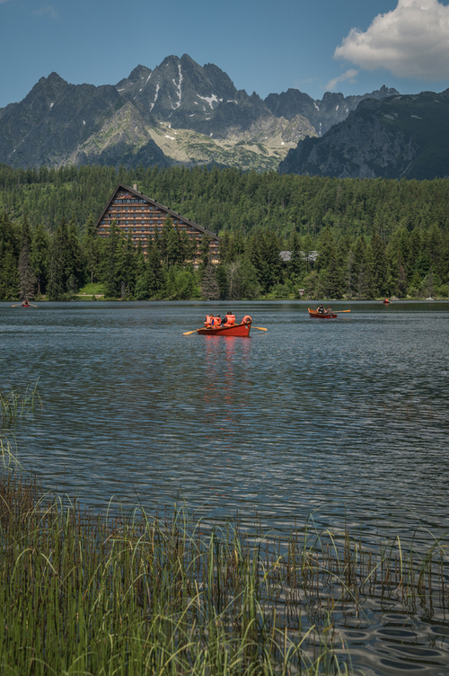 Urokliwe jezioro Štrbské Pleso -  miejscem spokoju i relaksu. Woda na jeziorze wydaje się być spokojna, jednak ten spokój zaburzają łodzie, malowane na czerwony kolor, dodają wyrazistego kontrastu do tej przyrodniczej scenerii. Ich jaskrawy odcień przyciąga uwagę i stanowi piękne uzupełnienie naturalnej palety barw tego miejsca. Nie tylko tafla wody ale i kolorystyka została zaburzona nadając dynamiki tego krajobrazu.

Światło słoneczne odbija się od wody, tworząc migoczące refleksy na powierzchni jeziora, co sprawia, że ​​cała sceneria wydaje się jeszcze bardziej magiczna. Ponadto, na horyzoncie ukazują się majestatyczne szczyty Tatr Wysokich, które dodają temu widokowi niepowtarzalnego uroku.

Wokół jeziora rozciąga się las, tworzący zieloną otulinę tego pięknego miejsca. Drzewa otaczające jezioro dodają uroku i zwiększają poczucie izolacji od zgiełku świata zewnętrznego.

Po drugiej stronie jeziora wyróżnia się wyjątkowy hotel o nietypowej i fascynującej architekturze. Ten hotel to prawdziwą ikoną tego miejsca. Jego architektura doskonale współgra z naturalnym pięknem okolicznej przyrody, tworząc niezapomniany widok. To magiczne miejsce nad Štrbské Pleso to idealne połączenie piękna natury, majestatycznych gór, kolorowych łodzi na jeziorze i fascynującej architektury, które pozostawi niezapomniane wrażenia każdemu, kto odwiedzi jezioro i jego okolicę.
