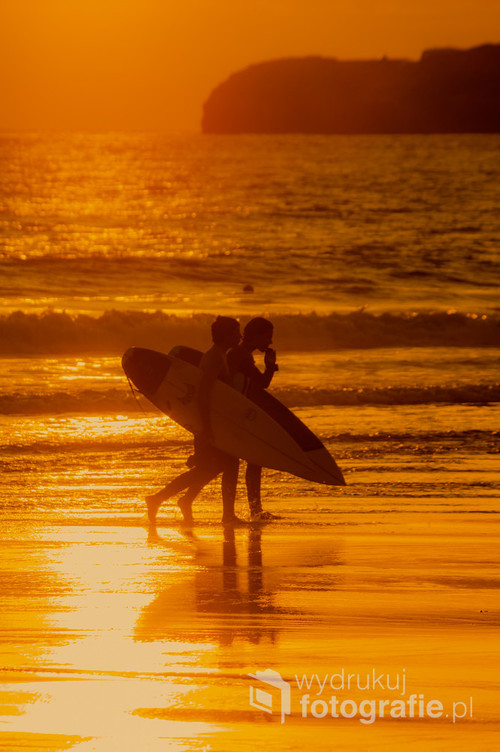 Dwóch surferów o zachodzie słońca, którzy właśnie wyszli z oceanu. W tle widoczny jest klif jednego z najpopularniejszych surferowych miasteczek w Portugalii, czyli Peniche gdzie dokładnie na tej plaży Supertubos odbywają się co roku jedne z największych na świecie mistrzostw World Surf League - coroczne tournee profesjonalnych zawodów surfingowych i transmisje każdego wydarzenia na żywo.
