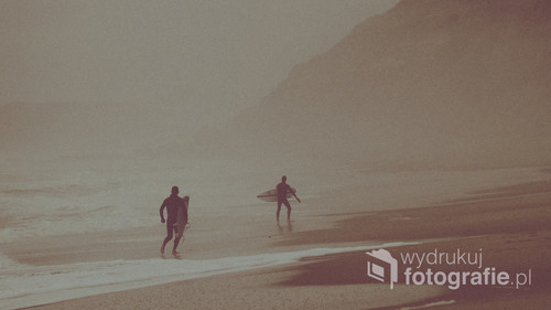Dwóch surferów o zachodzie słońca, na zamglonej plaży. Zdjęcie zrobione w trakcie opadu mgły oceanicznej, dzięki czemu powstał magiczny klimat.