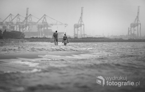 Fotografia zrobiona na Wyspie Sobieszewskiej (Ptasi Raj) Widok na parę idącą plażą w stronę Gdańska. Lekko wzburzone morze oraz delikatnie padająca mżawka.