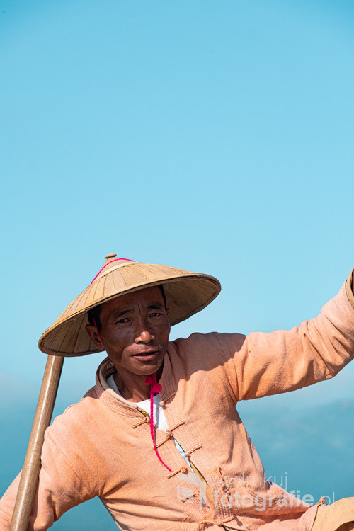 Spojrzenie rybaka na jeziorze Inle w dystrykcie Taunggyi (Mjanma, 2019). Kojarząca się z tańcem na wodzie sztuka łowiecka ludu Intha przetrwała po dziś dzień, jednakże przyszłość tej grupy etnicznej wydaje się mroczniejsza niż kiedykolwiek wcześniej. Narastające zagrożenie zaniku jeziora, który od wieków był źródłem podstawowych zasobów przeżycia, sięga powoli zenitu i tym samym końca pewnej ery dla rdzennych Intha. Na razie jednak pozostaje czekać i tańczyć na wodzie aż kolejna zdobycz wpadnie do rozstawionej siatki.