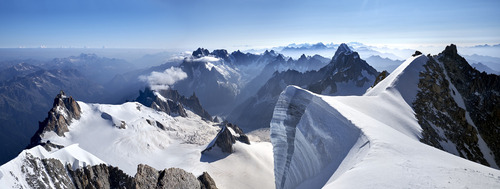 Fotografia wykonana na wierzchołku Mont Blanc du Tacul ukazuje nie tylko majestat gór, ale również kruchą ludzką obecność wobec ogromu przyrody. Widok na lodowiec Vallee Blanche, po którym wędrują ludzie, przypomina o nieustannym poszukiwaniu przez człowieka granic swoich możliwości. Aiguille du Midi po lewej stronie i schronisko Cosmique dodają kompozycji zdjęcia elementów ludzkiej ingerencji w krajobraz alpejski, stanowiąc punkty orientacyjne w tej imponującej przestrzeni.
Panorama rozciągająca się na horyzoncie, gdzie kolejne pasma gór wyłaniają się jedno po drugim, podkreśla nieskończoną wielowarstwowość i złożoność alpejskiego krajobrazu. Śnieżne granie, ukryte we mgle poranka, dodają scenerii tajemniczości i piękna, podczas gdy lodowe ściany budzące grozę przypominają o niebezpieczeństwach, które czyhają na tych, którzy odważą się na ich zdobycie.