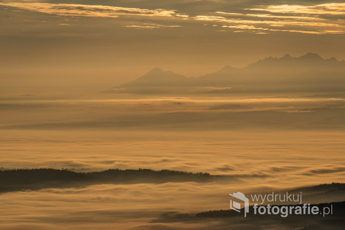 Widok na Tatry poprzez doliny górskie spowite mgłą, a wszystko w tonacji żółto-pomarańczowej.