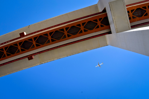 Zdjęcie zrobione w Lizbonie, wiszącego mostu na tle niebieskiego nieba. Widać lecący samolot 