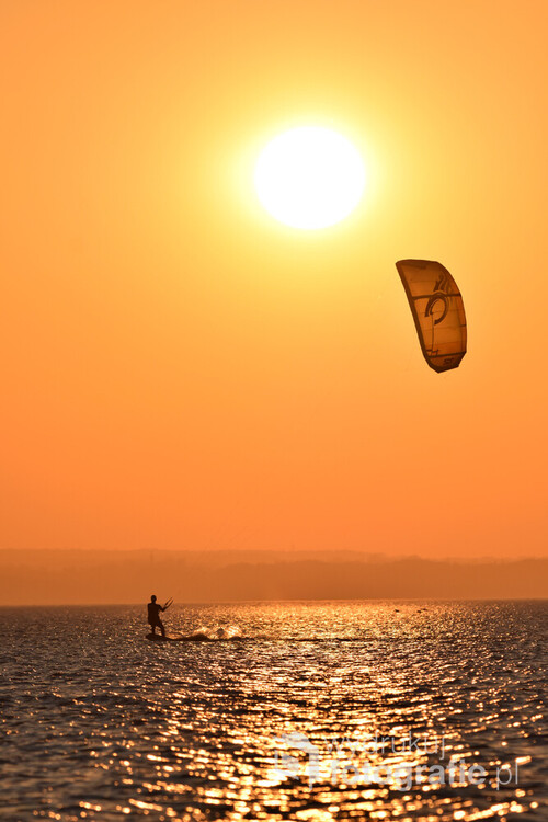 Fotografia przedstawia człowieka uprawiającego kitesurfing przy zachodzie słońca, Zdjęcie powstało nad morzem bałtyckim, w Juracie. 