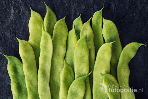 Green flat beans in pods lie on a stone slab. Top view.