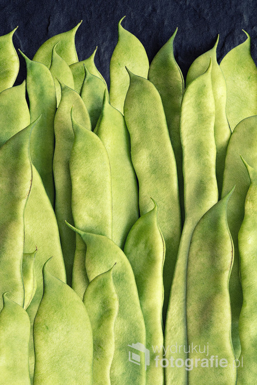 Green flat beans in pods lie on a stone slab. Top view.